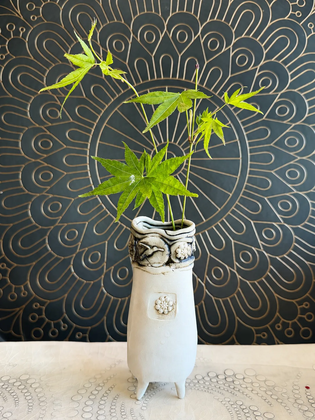Ikebana-style Japanese maple branch arrangement in a handmade textured ceramic vase against a dark patterned wall