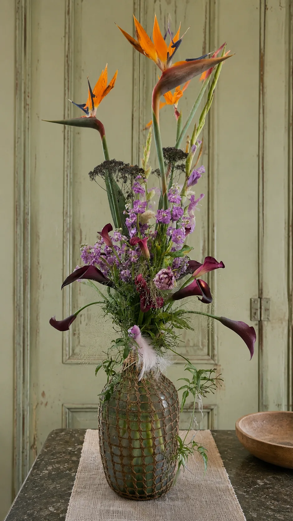 Bird of paradise and purple calla lily arrangement in a woven glass vase against a sage green door