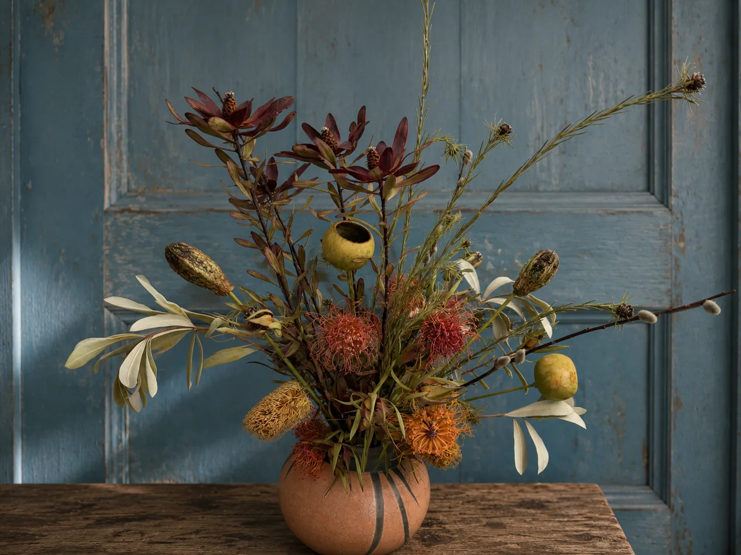 Dried protea and banksia arrangement in a terracotta vase against a weathered blue door