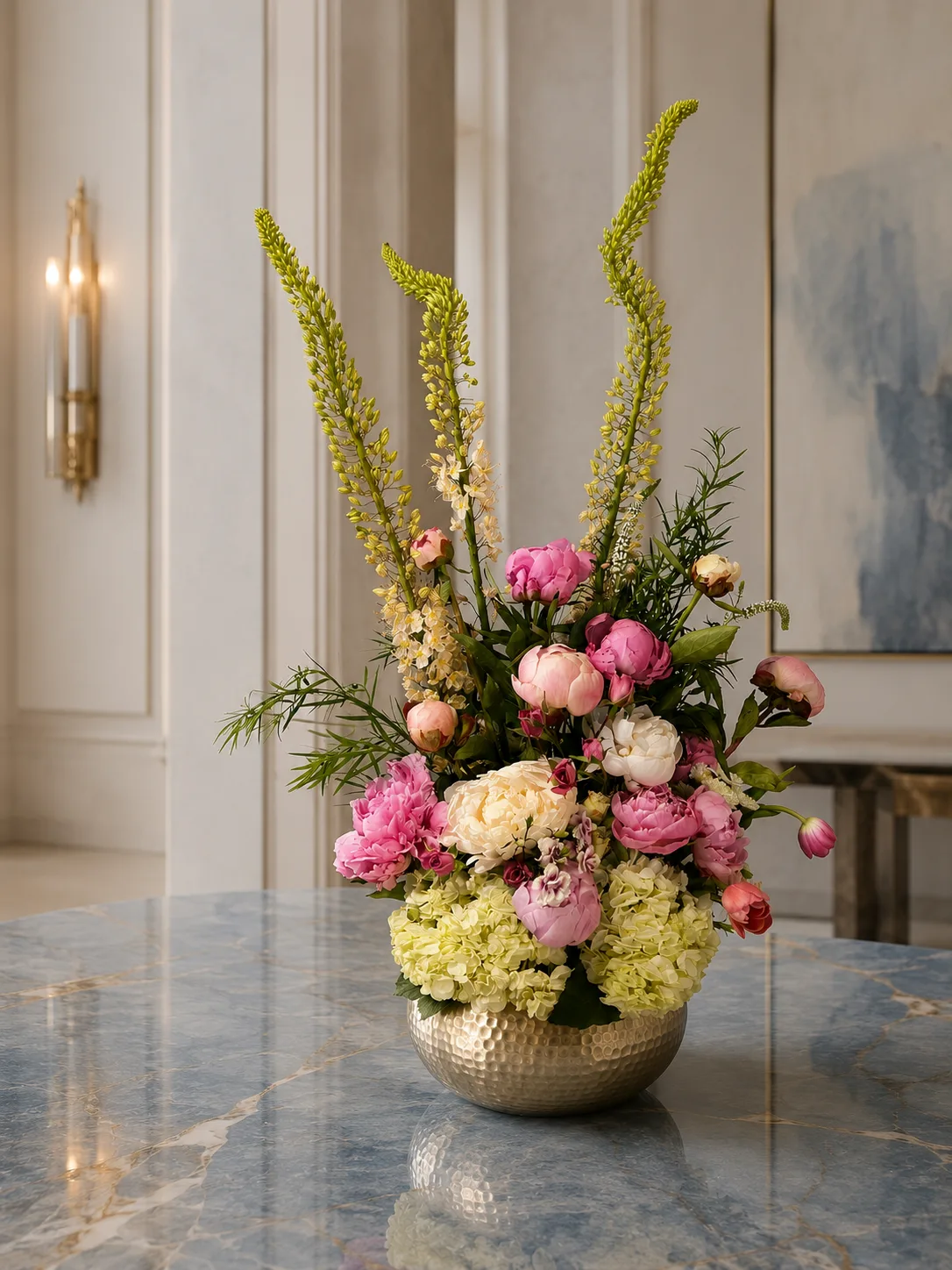Pink and cream peony arrangement with foxtail lily in a hammered gold bowl on a blue marble table