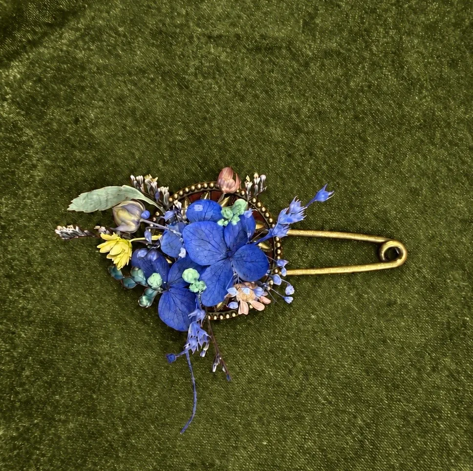 Dried blue hydrangea, yellow flower, and mixed wildflower brooch pinned to green velvet fabric with a gold safety pin