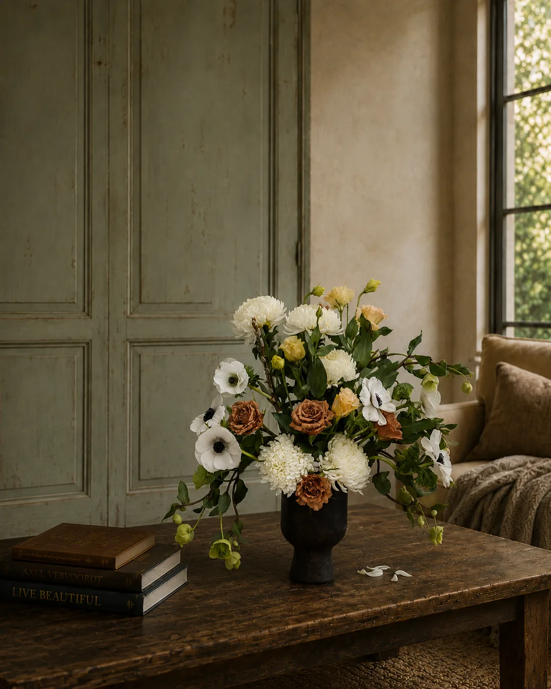 White chrysanthemum, anemone, and rust rose arrangement in a dark ceramic vase on a coffee table against teal paneling
