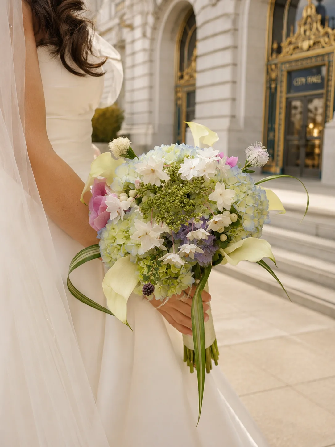 Bride holding a pastel hydrangea, dahlia, and calla lily bouquet with trailing ribbon outside San Francisco City Hall