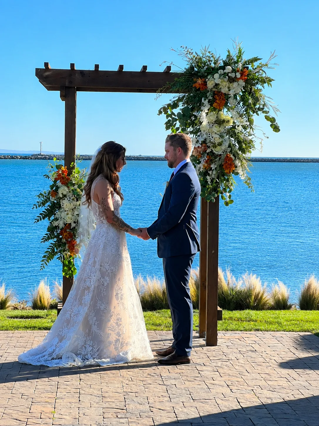 Bride and groom holding hands beneath a wooden ceremony arch dressed with a lush white and greenery floral arrangement, overlooking the water