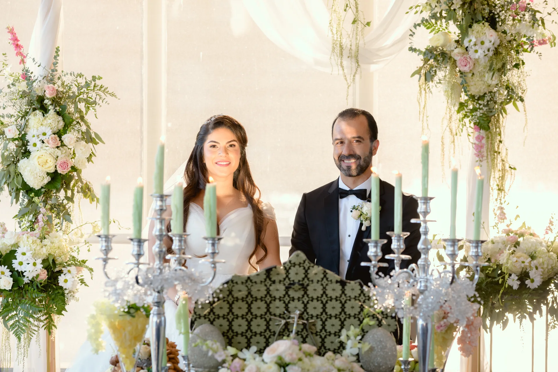 Bride and groom smiling at their Persian wedding ceremony, seated behind a sofreh aghd spread with candelabras and floral arrangements